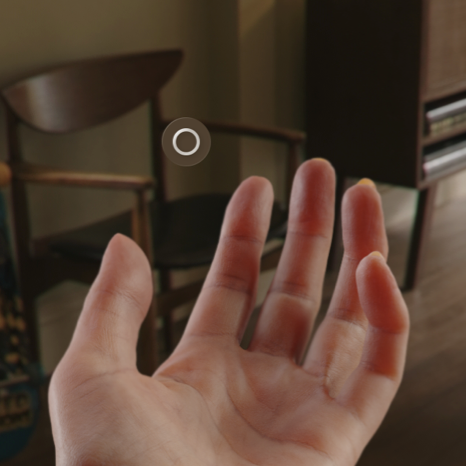 An image of a person's open hand with the palm facing upward, shown from the person's perspective. A button with a circle icon representing the Home indicator appears above the palm. The image background shows the room that's the person's surroundings.
